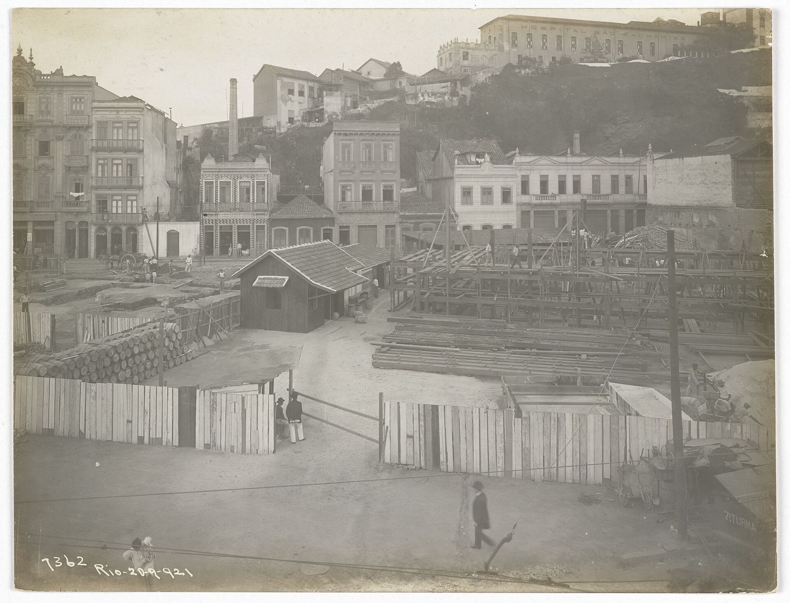 Fotografia antiga de um canteiro de obras com tapumes de madeira diante de sobrados no Rio de Janeiro.