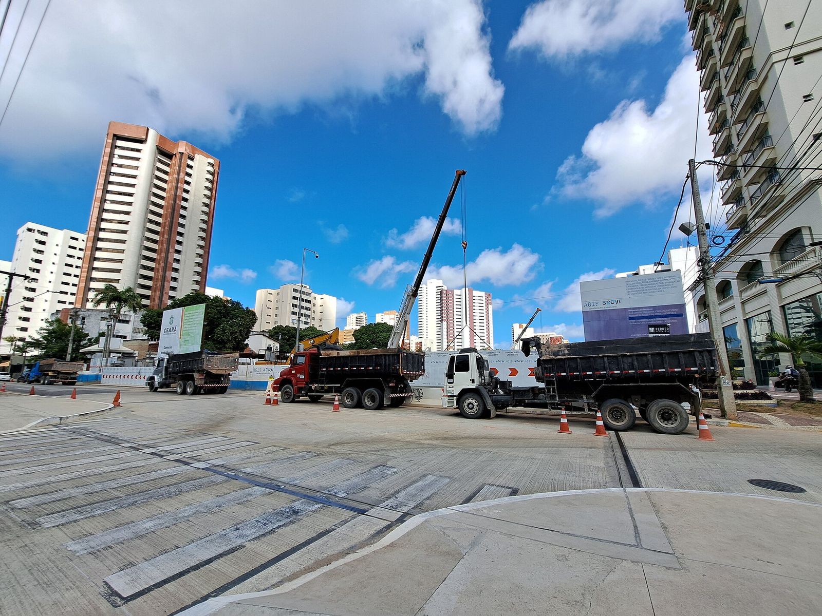 Obra de uma estação em avenida de Fortaleza, com caminhões e guindaste sob céu azul.