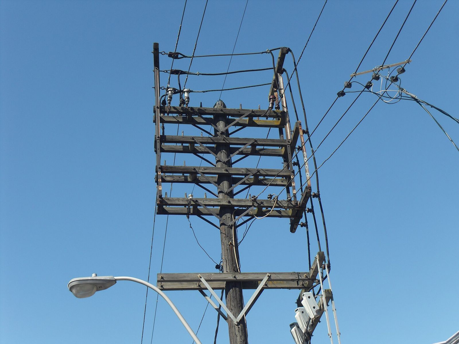 Poste de madeira com cruzetas, transformador e fios contra o céu azul.